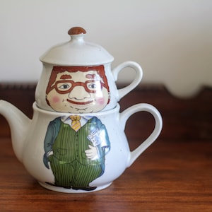 Può includere: Una teiera in ceramica bianca con la faccia di un uomo dei cartoni animati sul davanti. La teiera ha un coperchio marrone e un manico marrone. L'uomo indossa un abito verde e una cravatta.