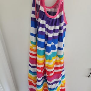 Könnte beinhalten: Ein bunt gestreiftes Sommerkleid mit Neckholder-Design. Das Kleid hat horizontale Streifen in Regenbogenfarben, darunter Rot, Orange, Gelb, Grün, Blau und Lila. Der Ausschnitt und die Träger sind rosa eingefasst.