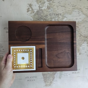 Puede incluir: Una bandeja de madera de nogal oscuro con una hendidura circular, un área rectangular empotrada y una ranura pequeña. Una baldosa cuadrada de cerámica con un diseño dorado y blanco está colocada en la bandeja. La bandeja es sostenida por una mano.