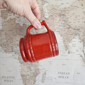 Op de afbeelding: Een rode keramische mok met een handvat en een textuurontwerp. De mok wordt in een hand gehouden tegen een wereldkaart achtergrond.