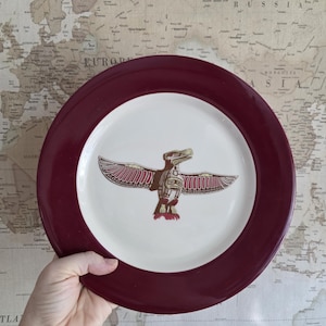 May include: A round, white plate with a maroon rim. The plate features a detailed, brown and red totem design in the center. The plate is held by a hand, with a world map in the background.