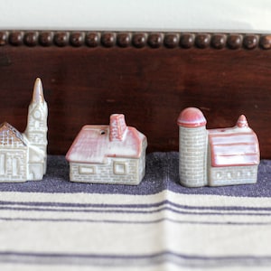 Op de afbeelding: Set van drie keramische miniatuurgebouwen: een kerk, een huis en een schuur. De kerk heeft een hoge toren, het huis een rood dak en de schuur een silo. Allemaal geschilderd in neutrale tinten met rode accenten.