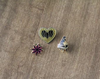 Puede incluir: Tres pines de esmalte sobre una superficie de madera. Los pines incluyen un corazón con detalles dorados, una estrella en rosa y morado, y un piano de cola en blanco y negro. Los pines son probablemente para una chaqueta o bolso.