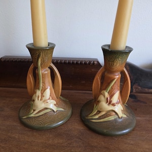 Puede incluir: Un par de candelabros de cerámica con velas altas de color crema. Los candelabros son marrones con diseños florales verdes y crema. La base es de color verde oscuro con un patrón texturizado. Los candelabros están sobre una superficie de madera.