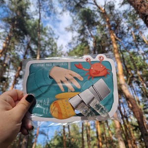 May include: A clear plastic package containing hair accessories is held up against a backdrop of trees. The package features a cartoon crab, a mitten, and the text "ИМАЙ КРАХА". The package also includes the text "@solemi.hair.accessories".