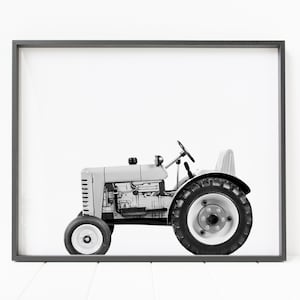 May include: A black and white photograph of a toy tractor in a gray frame. The tractor is a light gray color with black wheels and a black steering wheel.