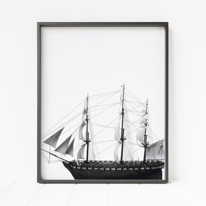 May include: A black and white photograph of a tall ship with sails billowing in the wind. The ship is framed in a black frame.