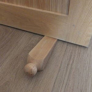 May include: A wooden doorstop with a rounded top and a rectangular base. The doorstop is made of light-colored wood and is resting on a wooden floor.