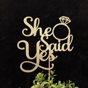 May include: Gold glitter cake topper with the words "She Said Yes" and a diamond ring.