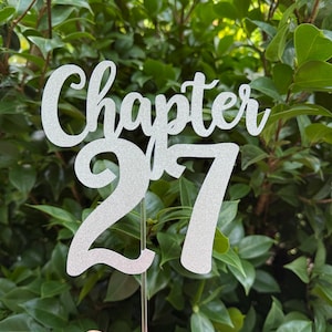 May include: A silver glitter cake topper with the words "Chapter 27" in a script font. The number 27 is in a bold, block font. The topper is held by a clear plastic stick, set against a backdrop of green leaves.