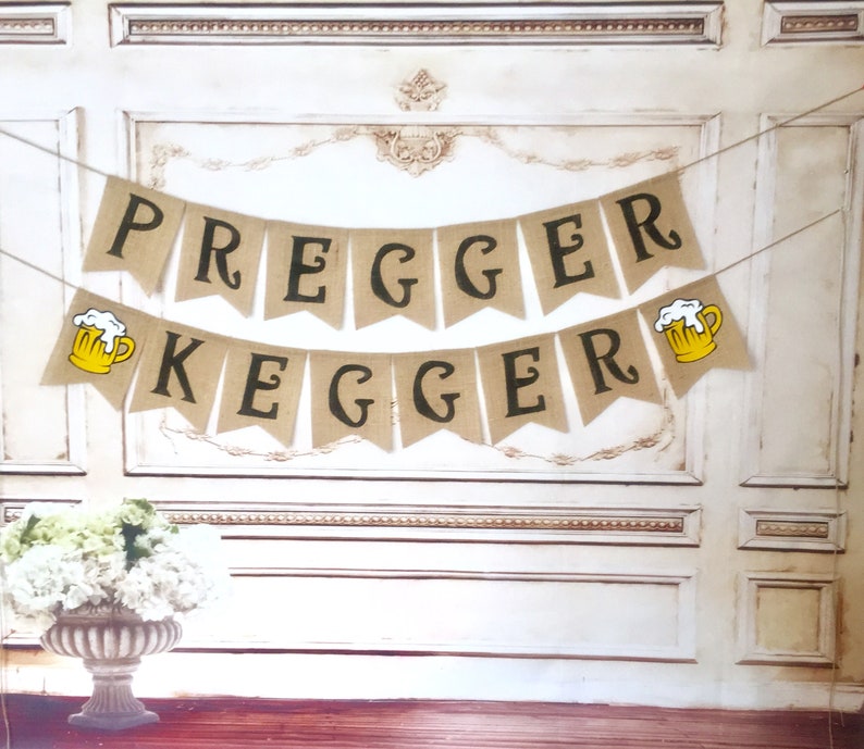 May include: A burlap banner with the text "Pregger Kegger" in black letters. The banner has two yellow beer mugs with white foam on top.