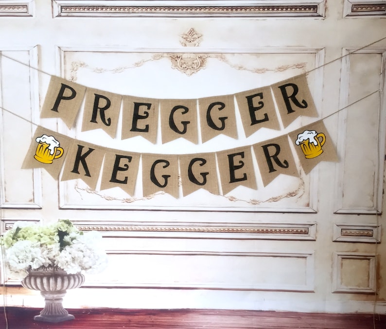 May include: A burlap banner with the text "Pregger Kegger" in black letters. The banner is decorated with two yellow beer mugs with white foam.