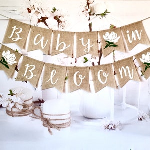 May include: A burlap banner with the words "Baby in Bloom" in white script. The banner is decorated with white flowers and green leaves.
