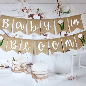 May include: A burlap banner with the words "Baby in Bloom" in white lettering. The banner is decorated with white tulips and green leaves.