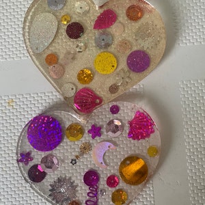 May include: Two heart-shaped resin coasters with a variety of colorful sequins and glitter embedded in the resin. The top coaster is light beige with pink, purple, gold, and silver sequins. The bottom coaster is clear with purple, gold, pink, and silver sequins.