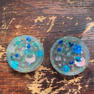 May include: Two round, clear resin coasters with a blue, white and pink glitter design. The coasters have a variety of shapes and sizes of glitter embedded in the resin.
