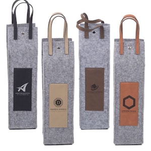 May include: Four gray felt wine bottle bags with different colored leather patches and logos. The logos are "Arkett Broadcasting", "Damen Jewels", "Ancient Kingdom", and "Lemdine Nexus".