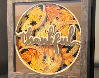 May include: A framed paper art piece with a circular design of layered paper leaves in shades of orange, yellow, and brown. The word "thankful" is written in brown paper letters in the center of the design.