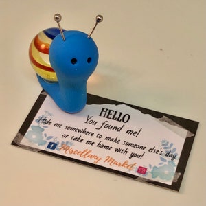 May include: A blue and yellow snail figurine with a message card that reads "HELLO You found me! Hide me somewhere to make someone else's day or take me home with you! f Miscellany Market"