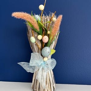 May include: A decorative Easter bouquet featuring speckled pastel eggs, faux pampas grass, and sprigs of greenery. The arrangement is tied with a light blue ribbon and set against a dark blue background.