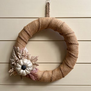 May include: A round wreath wrapped in burlap, adorned with a white pumpkin and fall foliage. The wreath is hanging on a light-colored wooden wall, creating a rustic autumn decoration.
