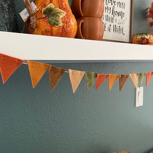 May include: A felt banner with triangular flags in shades of brown, orange, and green hangs on a white shelf. The flags are strung together with twine. The text "I love most my home & who I share it with." is visible on a framed sign behind the banner.