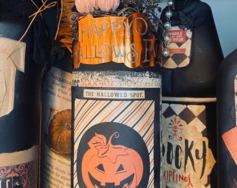 May include: A decorative glass bottle wrapped in orange twine at the neck, featuring a label with a jack-o'-lantern graphic, diagonal stripes, and the text "HAPPY HALLOWEEN" and "THE HALLOWED SPOT" printed in black and orange on a black background.