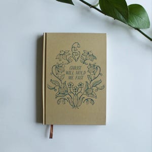 May include: A brown notebook with a blue floral design and the text "CHRIST WILL HOLD ME FAST".