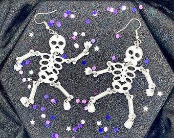May include: A pair of white skeleton earrings with silver hooks. The skeletons are posed in a dancing position. The earrings are on a black background with purple and white glitter.
