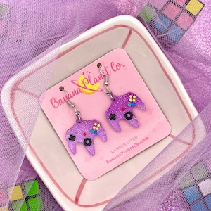 Peut inclure: Boucles d'oreilles en forme de manette de jeu avec des paillettes violettes et des crochets argentés. Les boucles d'oreilles sont sur un fond rose avec le texte "Banana Planet Co." sur la carte.