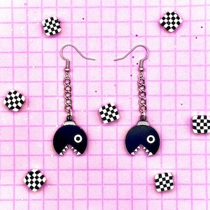 Może przedstawiać: Para czarno-białych kolczyków Pac-Mana z srebrnymi łańcuszkami. Kolczyki znajdują się na różowo-białym tle w kratkę.