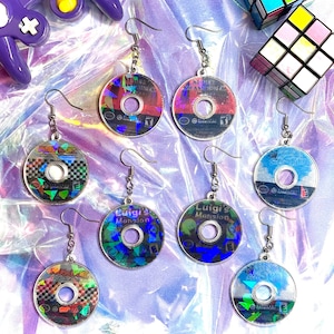 Könnte beinhalten: Acht Paar regenbogenfarbene Ohrringe mit Designs von Videospiel-Kassetten. Die Ohrringe sind silberfarben und zeigen verschiedene Spieltitel wie "Luigi's Mansion", "Super Mario Sunshine" und "Metroid Prime 2".