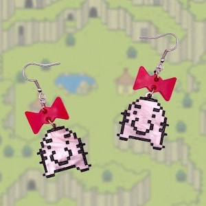 May include: A pair of earrings featuring a pixelated character with a red bow. The character is pink with black outlines and a smiling face. The earrings are made of polymer clay.