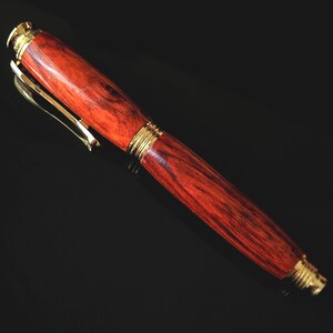 Dark shot of a handcrafted wooden fountain pen cap showing smooth reddish wood grain and gold clip on reflective black surface.