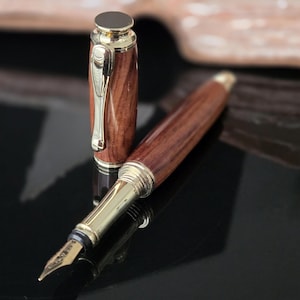 Close-up of handcrafted wooden fountain pen nib and cap with polished gold metal accents on glossy black surface.