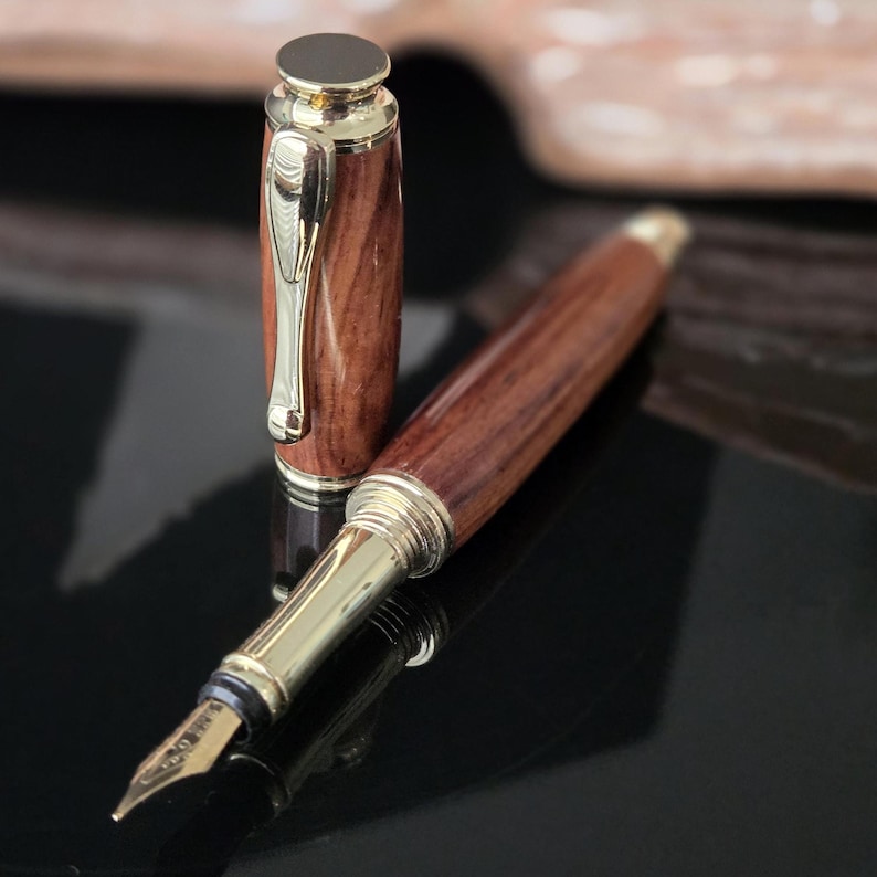 Close-up of handcrafted wooden fountain pen nib and cap with polished gold metal accents on glossy black surface.