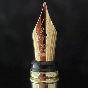 Detailed macro photograph of gold colored fountain pen nib with engraved decorative pattern and breather hole, shown against a black background