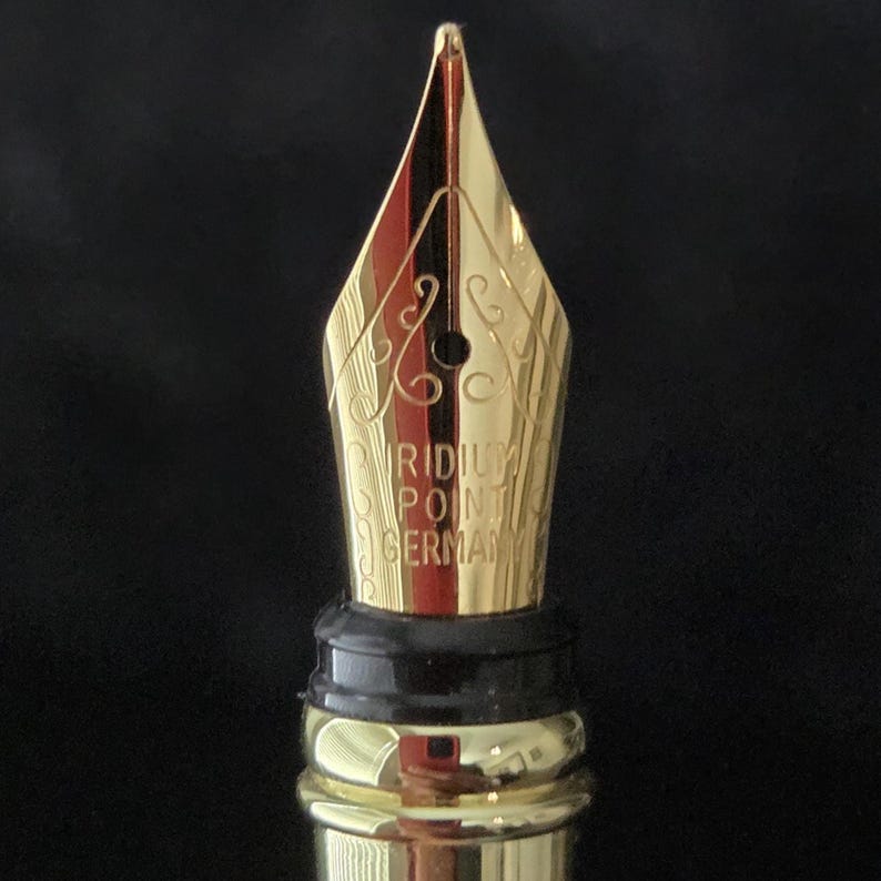 Detailed macro photograph of gold colored fountain pen nib with engraved decorative pattern and breather hole, shown against a black background