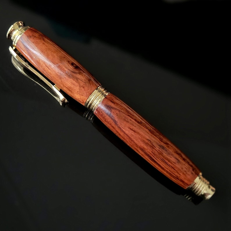Handcrafted wooden fountain pen cap showing smooth reddish wood grain and gold clip on reflective black surface.