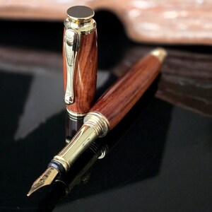 Luxury handmade wooden fountain pen with rich reddish grain and gold hardware resting on reflective black surface with open book behind.