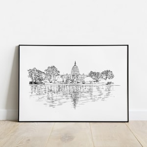 May include: Black and white sketch of the U.S. Capitol building, reflected in the water. The artwork is framed in a black border and features detailed linework of the building, trees, and water. The print is displayed on a white wall.