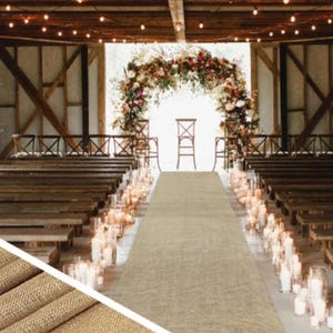 May include: A rustic wedding ceremony setup inside a barn. The aisle features a tan runner, flanked by candles in glass holders. A floral arch frames the altar, with wooden benches for guests. Overhead, string lights add a warm glow.