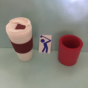 May include: A white travel coffee mug with a brown sleeve and lid, a red cup, and a blue golf-themed sticker on a light blue surface. The mug has a brown square on the lid.