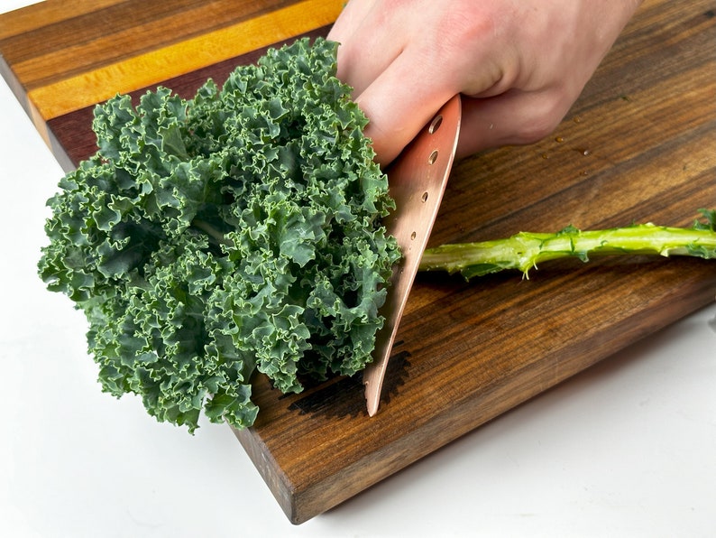 May include: A person is using a copper knife to remove the stem from a bunch of fresh green kale on a wooden cutting board.