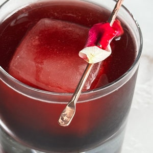 May include: A glass filled with a dark red liquid, a large ice cube, and a rose petal on a gold skewer.
