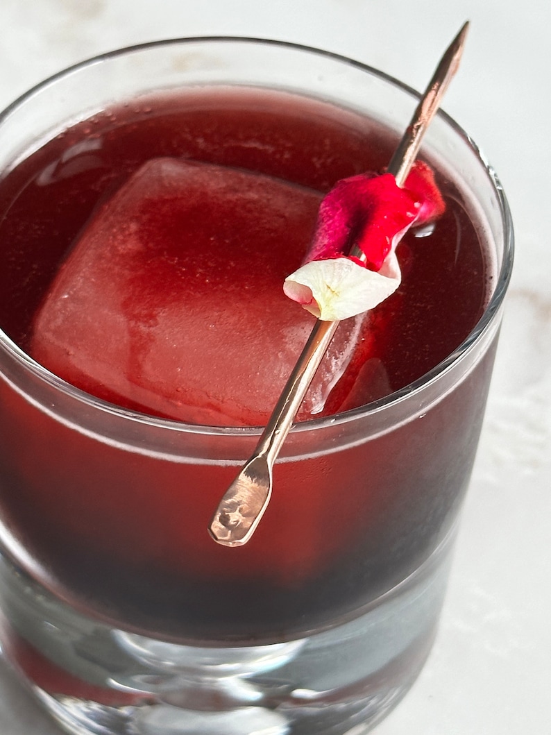 May include: A glass filled with a dark red liquid, a large ice cube, and a rose petal on a gold skewer.