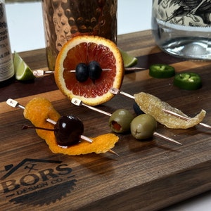 May include: A wooden cutting board with a logo that says "Bors Design Co."  There are five cocktail skewers with various garnishes, including a blood orange slice with blueberries, a cherry with an orange peel, two green olives, and a piece of candied ginger.