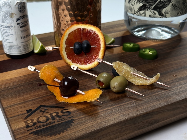May include: A wooden cutting board with a logo that says "Bors Design Co."  There are five cocktail skewers with various garnishes, including a blood orange slice with blueberries, a cherry with an orange peel, two green olives, and a piece of candied ginger.