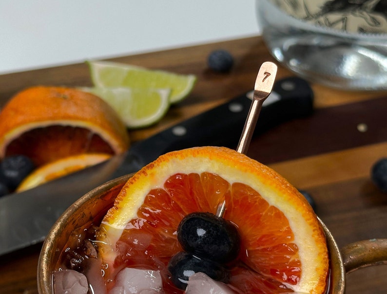 May include: A copper Moscow mule mug filled with ice, a dark red liquid, and a slice of blood orange. A copper stirring stick with the number "7" engraved on it is in the mug. There are blueberries and a slice of lime in the background.