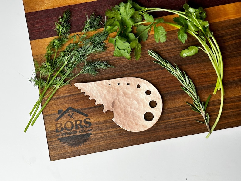 May include: A wooden herb stripper with a scalloped edge and multiple holes for stripping herbs. The stripper is on a wooden cutting board with sprigs of dill, cilantro, and rosemary.